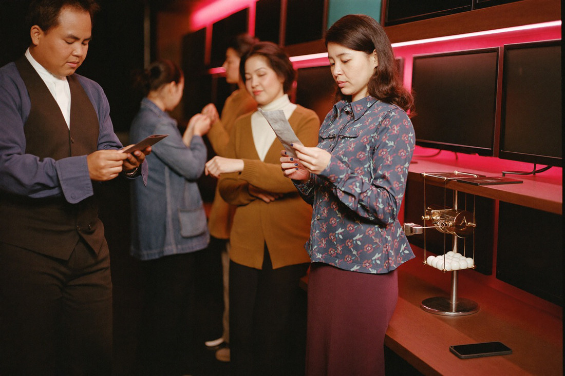 A clerk in a dark waistcoat handing a small folded paper slip across a counter ledge to a woman in a high necked blouse, a wire mesh drum cabinet behind him