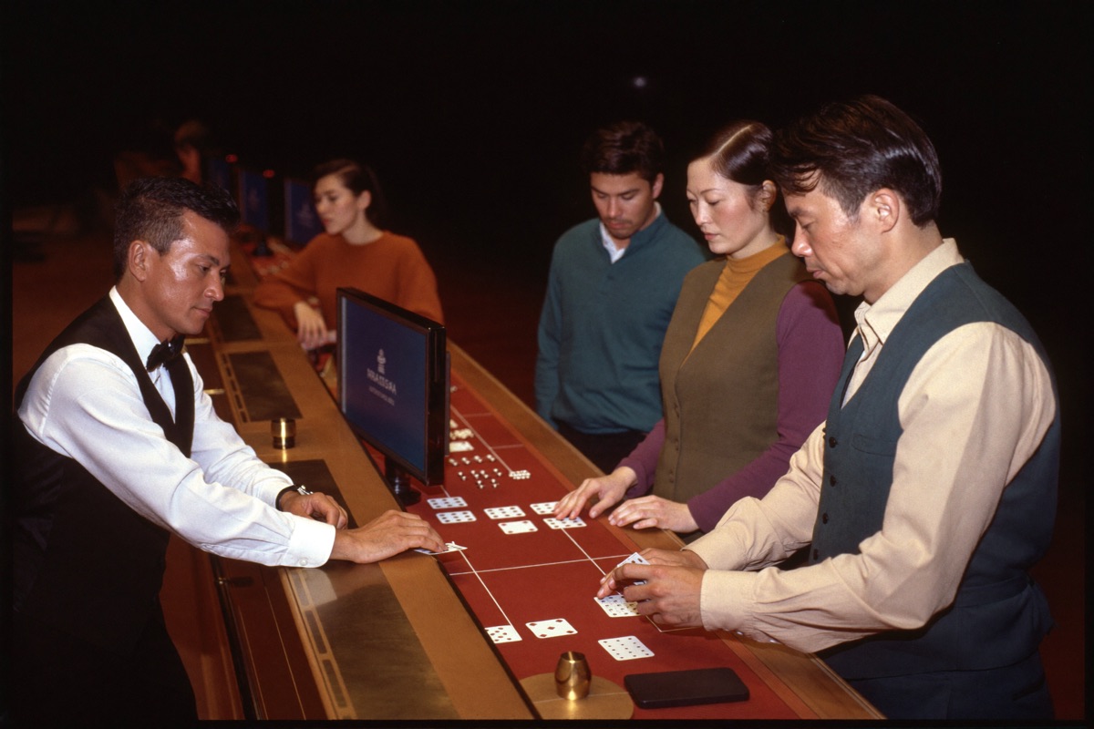 A dealer in a waistcoat standing where the card tables meet a bank of slot cabinets, turning back towards the felt while two seated players stay at the cabinets