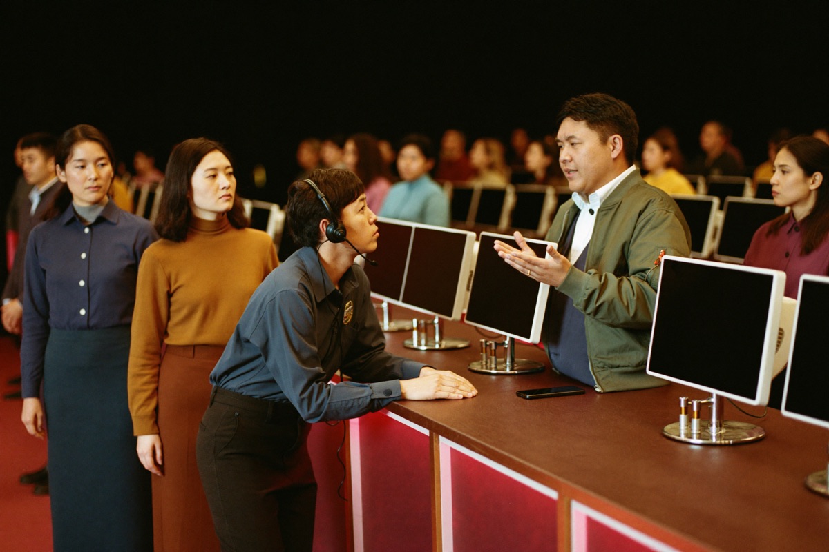 A host in a headset at a small service desk leaning across the counter to listen to a seated player in a buttoned shirt
