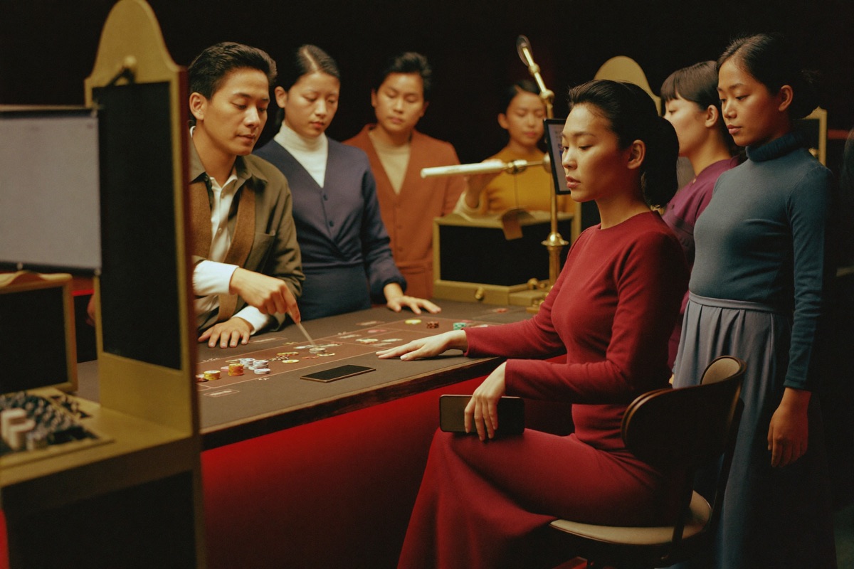 A woman still seated at a baccarat table holding a plain dark phone low beside the felt, her other hand on her chips while the dealer waits mid deal