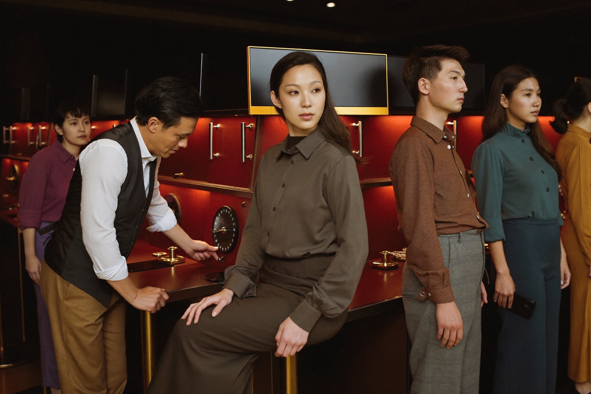 A young woman in a high necked blouse at her first slot cabinet with a host in a waistcoat crouched beside the stool pointing at the stake dial