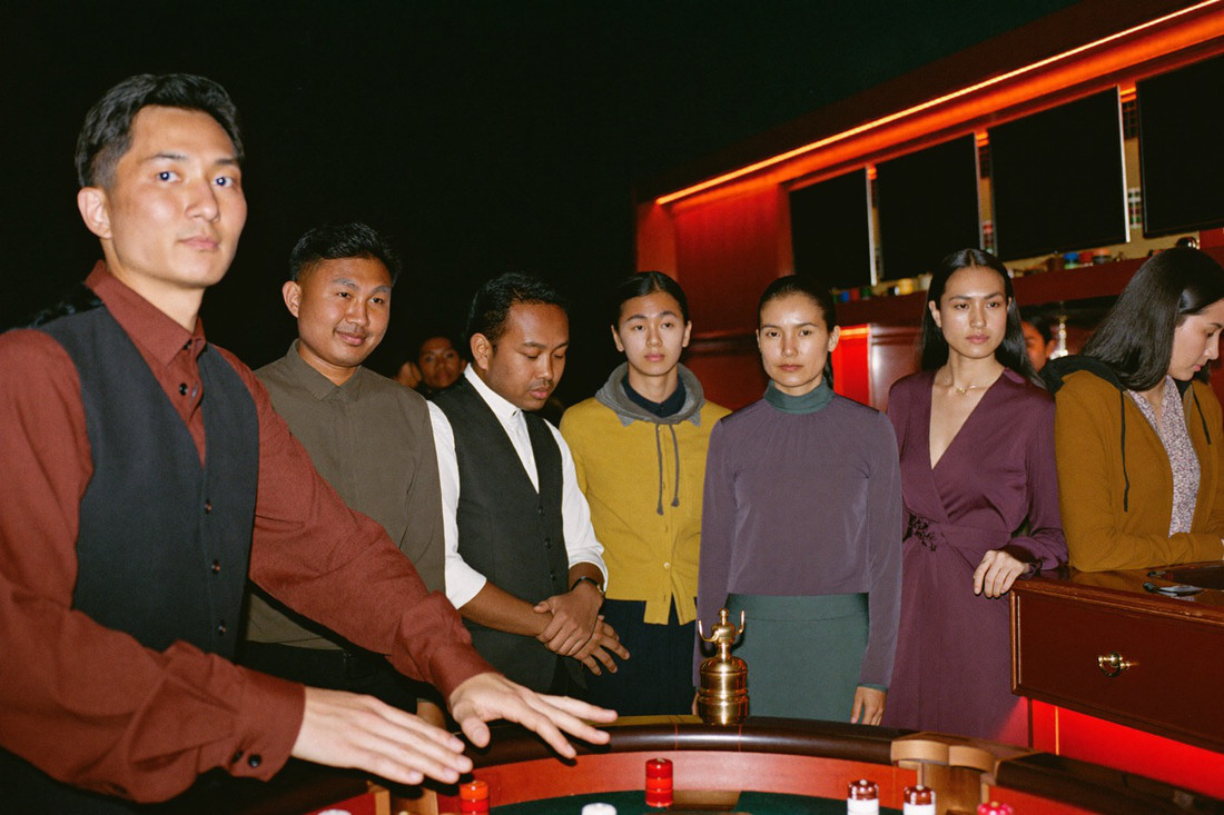 A roulette wheel in the foreground with the ball still running, the croupier in a waistcoat with one hand on the rim and four players standing at the rail