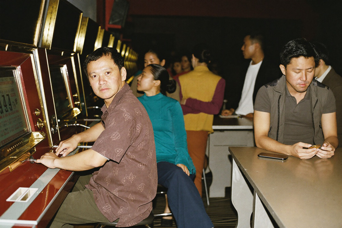 Three players at adjacent slot cabinets seen from behind, the nearest a woman in a dark blazer reaching for the stake button while the hall falls away into darkness
