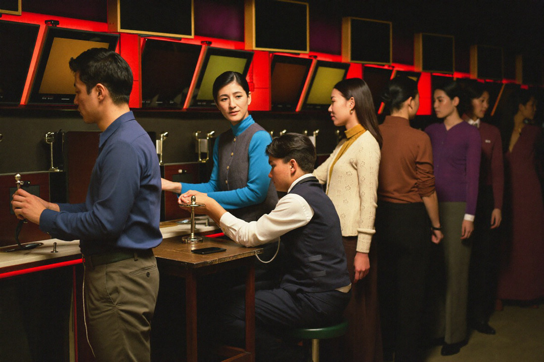 A Malaysian Malay man, a Chinese woman and an Indian man at three adjacent slot cabinets on a busy evening, a host in a dark uniform crouched beside the middle stool