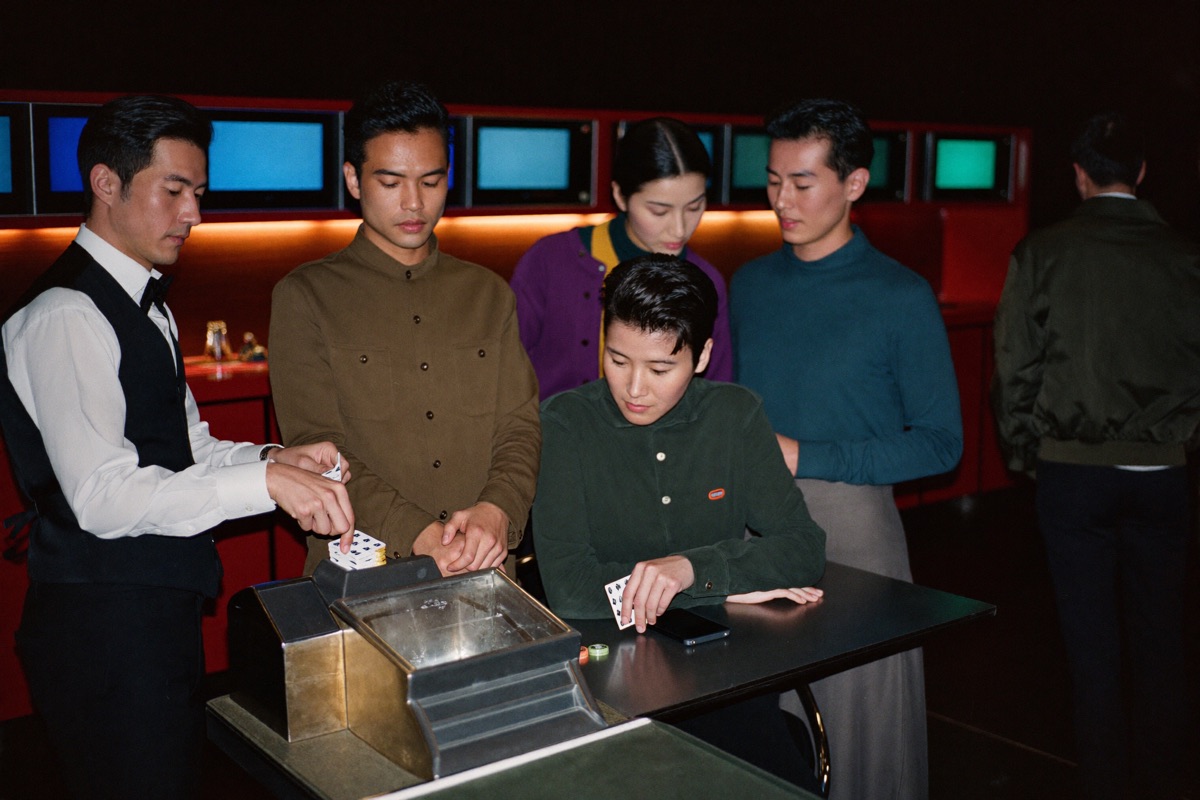 A live baccarat table from the player's seat, the dealer in a black waistcoat drawing a card from the shoe with both hands and a chip tray sharp on the felt