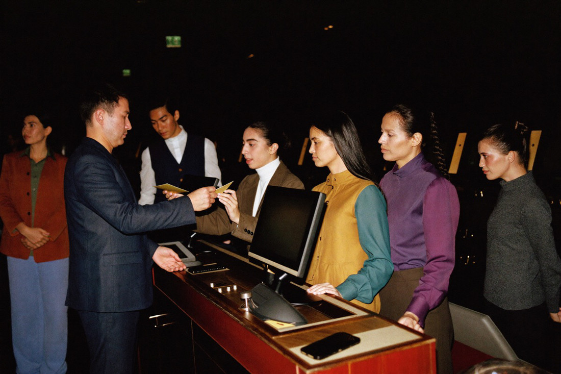 A host at a podium on the floor handing a small plain card to a player in a dinner jacket, two more players waiting behind him