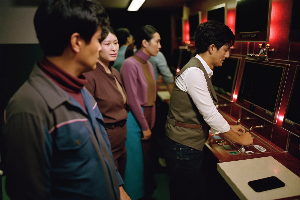 Close over the shoulder of a man in a buttoned shirt at a tall slot cabinet, five reels blurred mid spin and his thumb resting on the stake button
