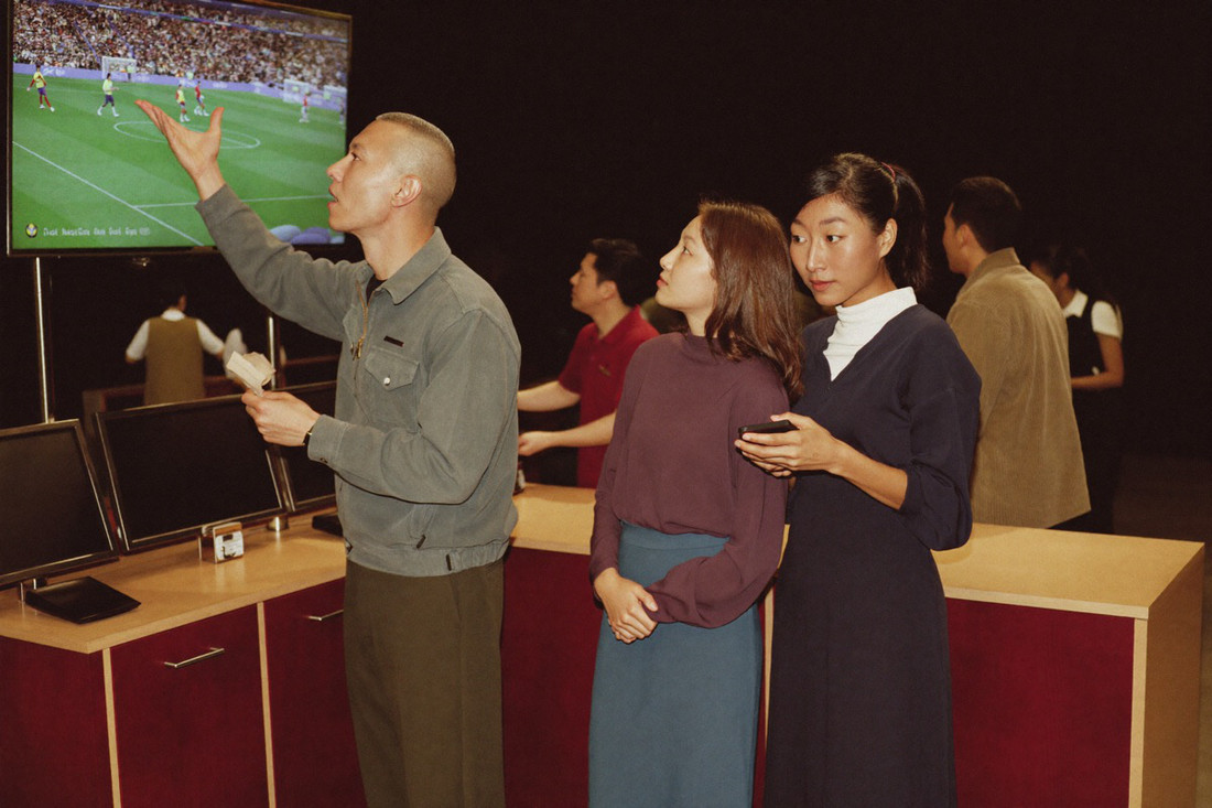 Three friends in plain shirts in the sports corner of a casino watching an association football match, one of them holding a folded paper slip
