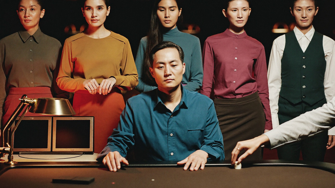 A single chip standing upright on the felt of an empty baccarat table under a low lamp, the dealer's hand entering from the right