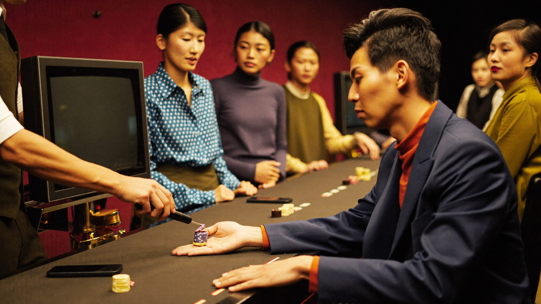 A croupier's rake drawing a short line of chips back along a baccarat layout towards a seated player, both pairs of hands in frame