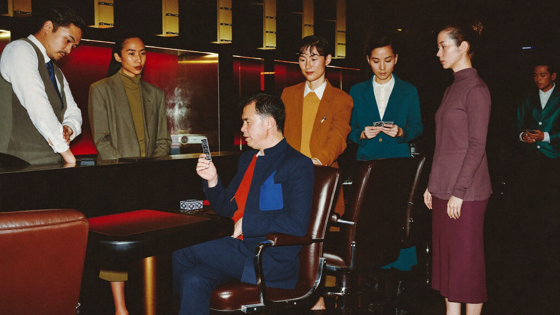 A high limit baccarat salon with four heavy leather chairs, one player in a dinner jacket seated and a dealer in a waistcoat standing at the shoe