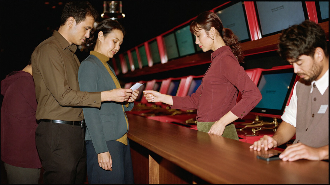 A couple at the member desk taking a small plain receipt from a clerk in a waistcoat, both in buttoned shirts