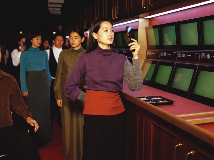 A pair of hands holding a plain dark phone in portrait at the rail of a live table, the dealer's waistcoat and the chip tray soft behind it