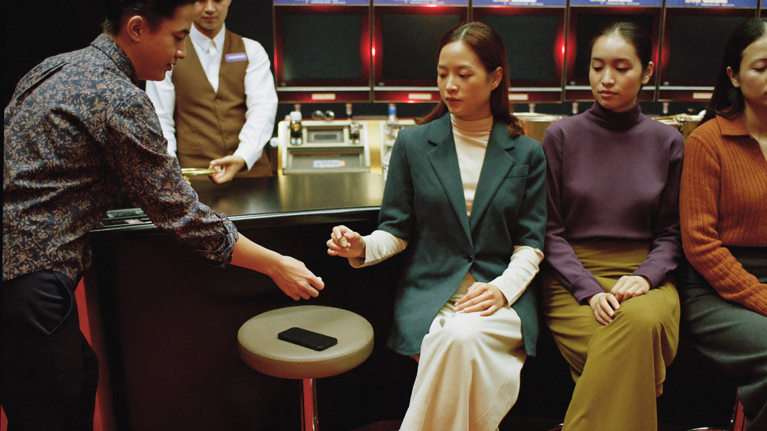 A host in a waistcoat setting a single small chip down in front of a woman in a high necked blouse at an empty slot stool