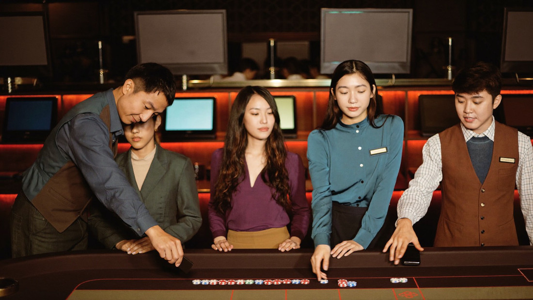 A pit clerk in a waistcoat sweeping a short row of chips back towards a seated player across the baccarat felt